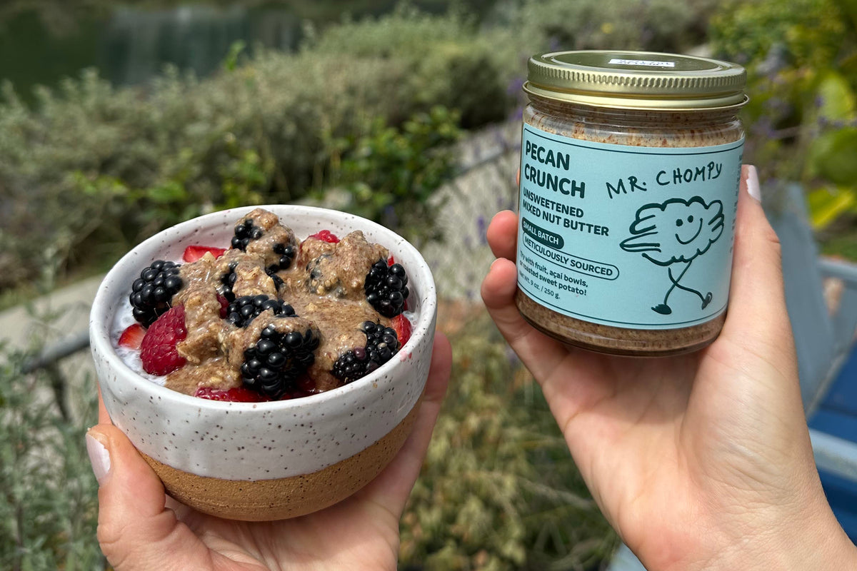 Two hands hold a small jar of Mr. Chown's Pecan Crunch spread and a bowl of the spread topped with berries, with a blurred outdoor canal and houses in the background.